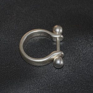 May include: A silver-colored shackle ring with a rounded D-shape and a removable pin. The ring has a smooth, slightly aged appearance. The ring is a piece of jewelry.