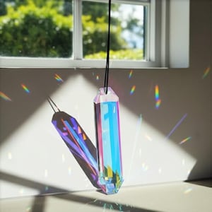 May include: A prism-shaped crystal suncatcher hanging from a black cord. The crystal reflects light, creating a rainbow effect on the wall. The crystal is a mix of pink, blue, and clear colors. The background includes a window and greenery.