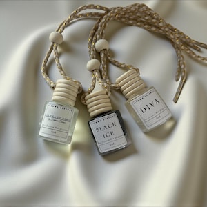 May include: Three small, clear glass bottles with wooden caps and braided gold cords hang against a white background. The bottles are labeled "DIVA", "BLACK ICE", and "FIAMMA DE PARIS". The bottles are for scented car diffusers.