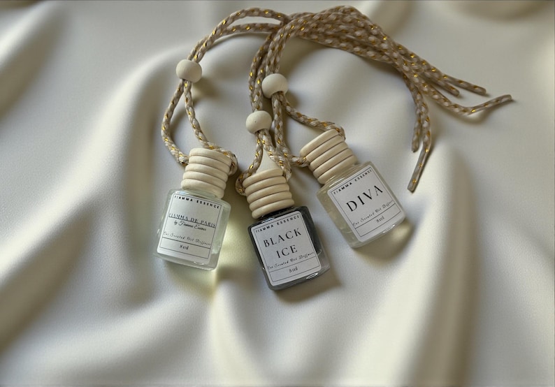 May include: Three small, clear glass bottles with wooden caps and braided gold cords hang against a white background. The bottles are labeled "DIVA", "BLACK ICE", and "FIAMMA DE PARIS". The bottles are for scented car diffusers.