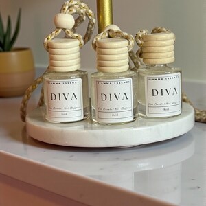 May include: Three small, clear glass bottles with wooden caps and hanging cords, labeled "DIVA" and "Basil." The bottles sit on a white marble tray. The bottles are filled with a clear liquid. The text "FIAMMA ESSENCE" is above the word "DIVA".