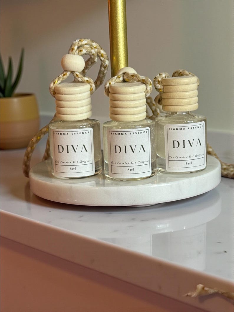 May include: Three small, clear glass bottles with wooden caps and hanging cords, labeled "DIVA" and "Basil." The bottles sit on a white marble tray. The bottles are filled with a clear liquid. The text "FIAMMA ESSENCE" is above the word "DIVA".