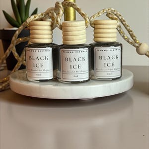 May include: Three small glass bottles of "Black Ice" scented oil diffusers, each with a wooden cap and a hanging string. The bottles are displayed on a white, round platform. The text "FIAMMA ESSENCE" and "8 ml" are visible.