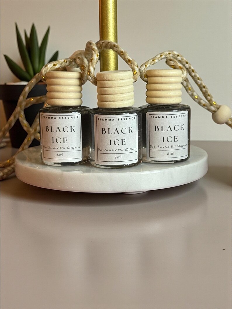 May include: Three small glass bottles of "Black Ice" scented oil diffusers, each with a wooden cap and a hanging string. The bottles are displayed on a white, round platform. The text "FIAMMA ESSENCE" and "8 ml" are visible.