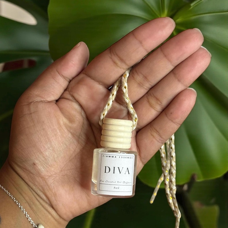 May include: A small, clear glass bottle with a wooden cap and a braided cord. The label reads "DIVA" and "Fiamma Essence". The bottle is held in a hand against a green leafy background. The product is an air freshener.