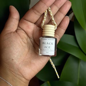 May include: A small, clear glass bottle with a wooden cap and a gold and white string. The label reads "BLACK ICE" and "For Scented Oil Diffuser". The bottle is held in a hand, with a silver bracelet visible in the foreground.