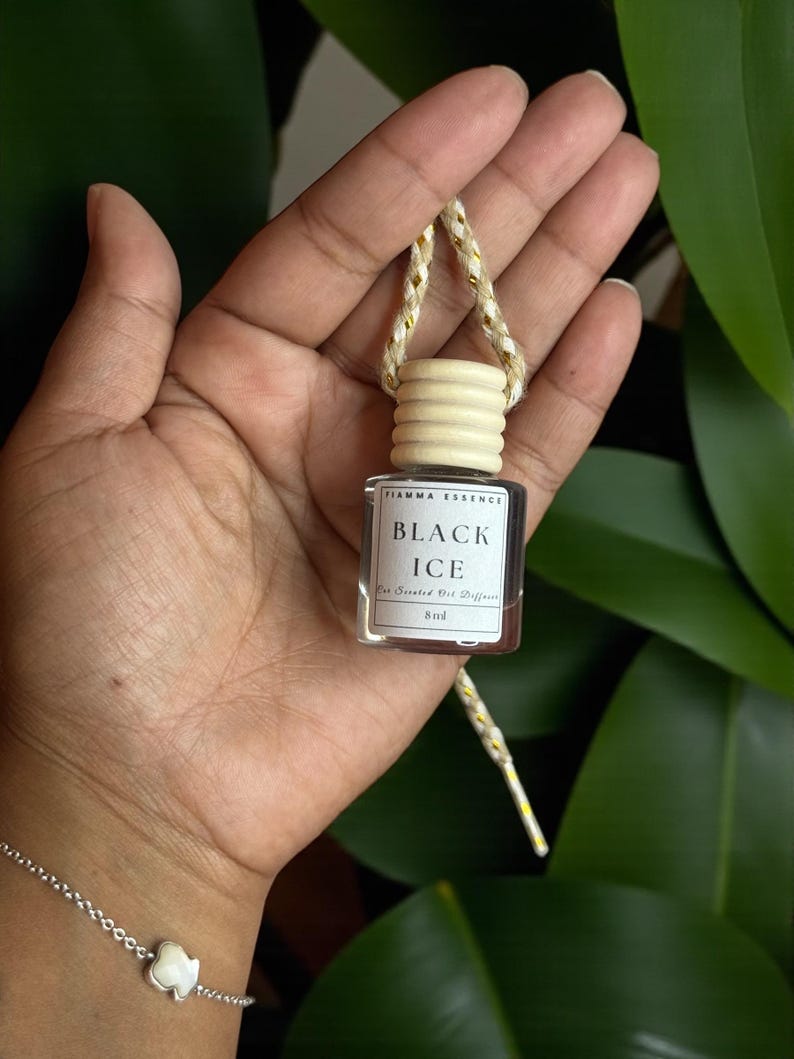 May include: A small, clear glass bottle with a wooden cap and a gold and white string. The label reads "BLACK ICE" and "For Scented Oil Diffuser". The bottle is held in a hand, with a silver bracelet visible in the foreground.