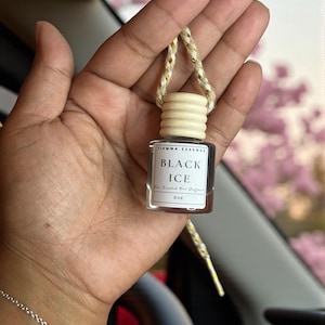 May include: A small, rectangular glass bottle with a wooden cap and a gold and white string, labeled "BLACK ICE" and "Fiamma Essence." The bottle is held in a hand, with a silver bracelet visible. The background is blurred.