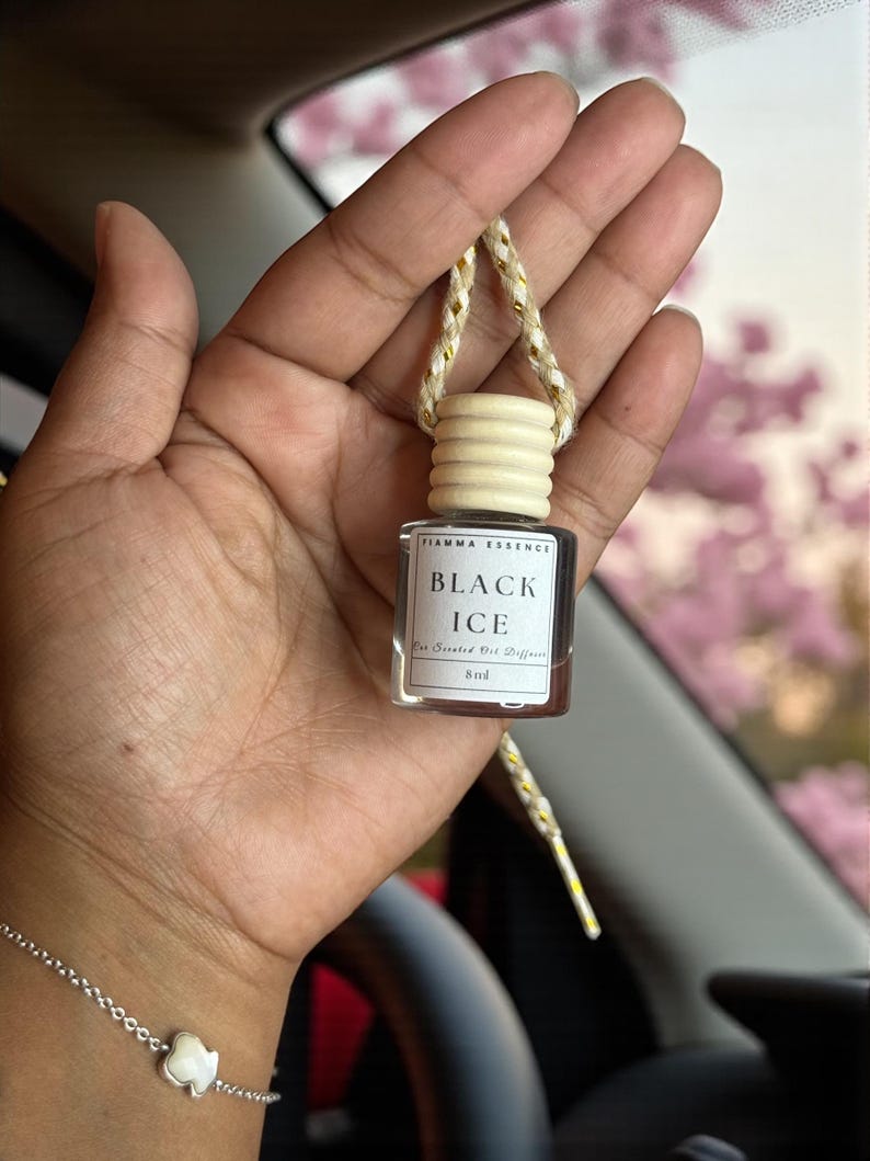 May include: A small, rectangular glass bottle with a wooden cap and a gold and white string, labeled "BLACK ICE" and "Fiamma Essence." The bottle is held in a hand, with a silver bracelet visible. The background is blurred.