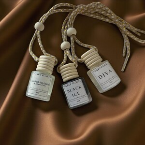May include: Three small, clear glass bottles with wooden caps and hanging cords. Each bottle has a label with text, including "DIVA" and "BLACK ICE." The bottles are on a brown satin surface.