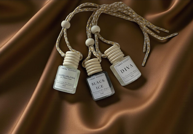 May include: Three small, clear glass bottles with wooden caps and hanging cords. Each bottle has a label with text, including "DIVA" and "BLACK ICE." The bottles are on a brown satin surface.