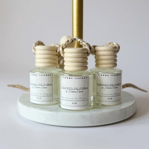 May include: Three small, clear glass bottles with wooden caps and braided cords, labeled "FIANNA DE PARIS" and "8 ml." The bottles sit on a white marble-like circular base, with a gold-colored stand in the background. The product is a car air freshener.