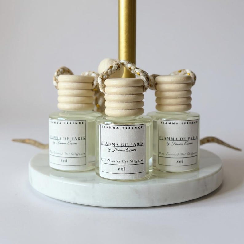 May include: Three small, clear glass bottles with wooden caps and braided cords, labeled "FIANNA DE PARIS" and "8 ml." The bottles sit on a white marble-like circular base, with a gold-colored stand in the background. The product is a car air freshener.