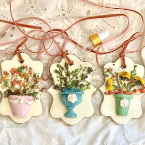 May include: Three decorative floral ornaments with dried flowers in pastel-colored pots. Each ornament is cream-colored with a scalloped edge and a brown ribbon for hanging. The pots are pink, blue, and green. A glass dropper is visible.