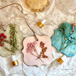May include: Three scalloped-edge hanging plaques in sage green, soft pink, and bright turquoise, each embedded with dried flowers and attached to a velvet ribbon. Two small clear glass dropper bottles with gold collars are placed nearby.
