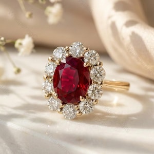 May include: A gold ring with a large, oval, deep red gemstone encircled by a halo of smaller, round, clear gemstones. The ring is displayed on a white surface with soft lighting, highlighting its sparkle. White flowers and cream fabric are in the background.