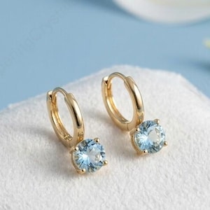 Puede incluir: Pendientes de aro dorados con piedras preciosas redondas de color azul claro. Los pendientes se exhiben sobre una superficie blanca texturizada contra un fondo azul claro. Pétalos de flores blancas dispersos en el fondo.