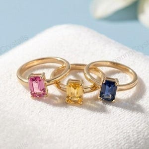 Peut inclure: Trois bagues en or avec des pierres rectangulaires roses, jaunes et bleues sont présentées sur un coussin de velours blanc. Une fleur blanche avec un centre jaune est floue en arrière-plan. Les bagues sont au centre.