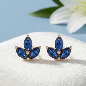 Peut inclure: Une paire de boucles d'oreilles à clous composée de trois pierres bleues taille marquise disposées en grappe en forme de feuille, serties dans une monture en métal doré. Les boucles d'oreilles sont présentées sur une surface en velours blanc.