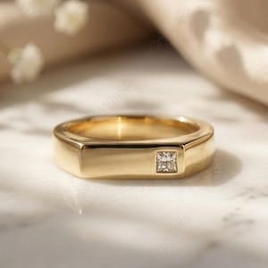 May include: A gold-colored ring with a square-cut diamond set in the center. The ring has a rectangular band and a polished finish. The background features soft, neutral tones and delicate white flowers.