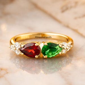 May include: A gold ring featuring a red and a green pear-shaped gemstone, flanked by small white gemstones. The ring is set on a marble surface, with the text "WhisperingCrystals" visible.