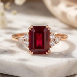 May include: A rose gold ring featuring a large, rectangular, deep red gemstone as the focal point. The ring is flanked by clusters of smaller, round, clear gemstones on each side. The setting is on a white marble surface.
