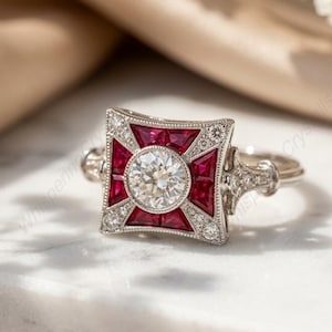 May include: A vintage ring with a square, ruby-red and diamond-studded centrepiece. The ring features a central, round, clear diamond, surrounded by smaller diamonds and red gemstones. The band is silver-toned, and the ring is set on a white marble surface.