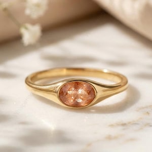 Op de afbeelding: Een gouden ring met een ovale, perzik-kleurige edelsteen. De ring ligt op een wit marmeren oppervlak, met een zachte, neutrale achtergrond. Delicate witte bloemen bevinden zich in de linkerbovenhoek.
