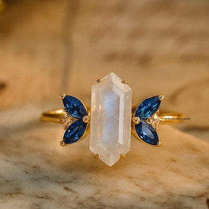 Könnte beinhalten: Ein goldener Ring mit einem großen, rechteckigen, irisierenden Mondstein-Mittelstein, flankiert von zwei Clustern aus drei blauen Edelsteinen. Das Design des Rings ist auf einer Marmoroberfläche platziert.