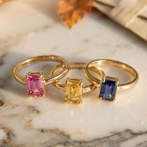 Könnte beinhalten: Drei goldene Ringe mit farbigen rechteckigen Edelsteinen: rosa, gelb und blau. Die Ringe werden auf einer Marmoroberfläche präsentiert, mit einem Vintage-Spiegel und Schreibwaren im Hintergrund. Die Ringe sind von Whispering Crystals.