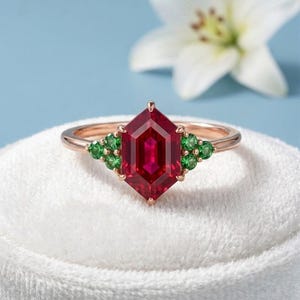 Könnte beinhalten: Ein roségoldener Ring mit einem großen, facettierten, sechseckigen Rubin-Mittelstein. Akzentuiert mit Clustern aus kleinen grünen Edelsteinen auf beiden Seiten. Der Ring ist vor einem weißen Hintergrund mit blauem Hintergrund platziert.