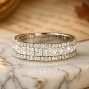 Może przedstawiać: Srebrny pierścionek z dwoma rzędami kwadratowych diamentów i rzędem mniejszych okrągłych diamentów. Pierścionek leży na marmurowej powierzchni, ukazując swój blask i skomplikowany design. To biżuteria.