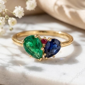 May include: A gold ring featuring a pear-shaped emerald, a pear-shaped sapphire, and a small ruby. The gemstones are set close together, creating a cluster design. The ring is displayed on a white surface.