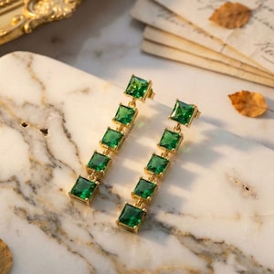 May include: A pair of gold-toned earrings with a vertical design. Each earring features five square, emerald-green gemstones. The earrings are displayed on a marble surface, with a gold-framed mirror and vintage papers in the background.