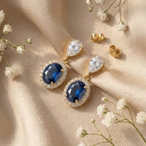 May include: Gold drop earrings with a large, oval, dark blue gemstone surrounded by small, clear stones. The earrings also feature a pear-shaped, clear gemstone at the top. The earrings are displayed on a gold-coloured fabric.