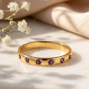 May include: A gold ring with a hammered finish, featuring small, star-shaped purple gemstones. The ring is displayed on a marble surface, with white flowers in the background. The ring is a piece of jewellery.