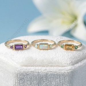 Könnte beinhalten: Drei goldene Ringe mit rechteckigen Edelsteinen. Die Ringe zeigen einen Amethyst, einen Opal und einen Citrin, jeweils flankiert von kleineren Akzentsteinen. Sie werden auf einer weißen, strukturierten Oberfläche vor einem hellblauen Hintergrund präsentiert.