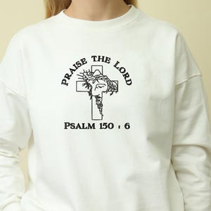 May include: White crew neck sweatshirt featuring the text "PRAISE THE LORD" above a cross graphic. The cross graphic has a detailed illustration and the text "PSALM 150:6" below it. The sweatshirt is a plain white colour.