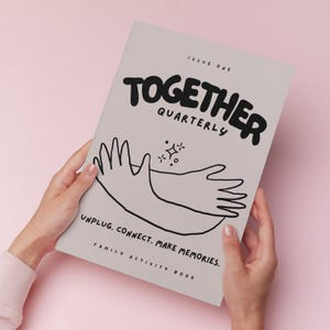 May include: A light gray "Together Quarterly" family activity book held by hands against a pink background. The cover features black text and a line drawing of hands. The text reads "Issue One", "Together Quarterly", and "Unplug. Connect. Make Memories."