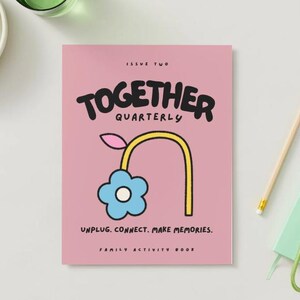 May include: A pink "TOGETHER QUARTERLY" family activity book with a blue flower illustration and the text "UNPLUG. CONNECT. MAKE MEMORIES." A pencil, a green notebook, and a bunch of grapes are also visible.