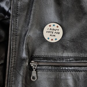 May include: A round, off-white button with the text "I DIDN'T VOTE FOR HIM" in black. The button is decorated with red, white, and blue stars. The button is pinned to a black leather jacket.