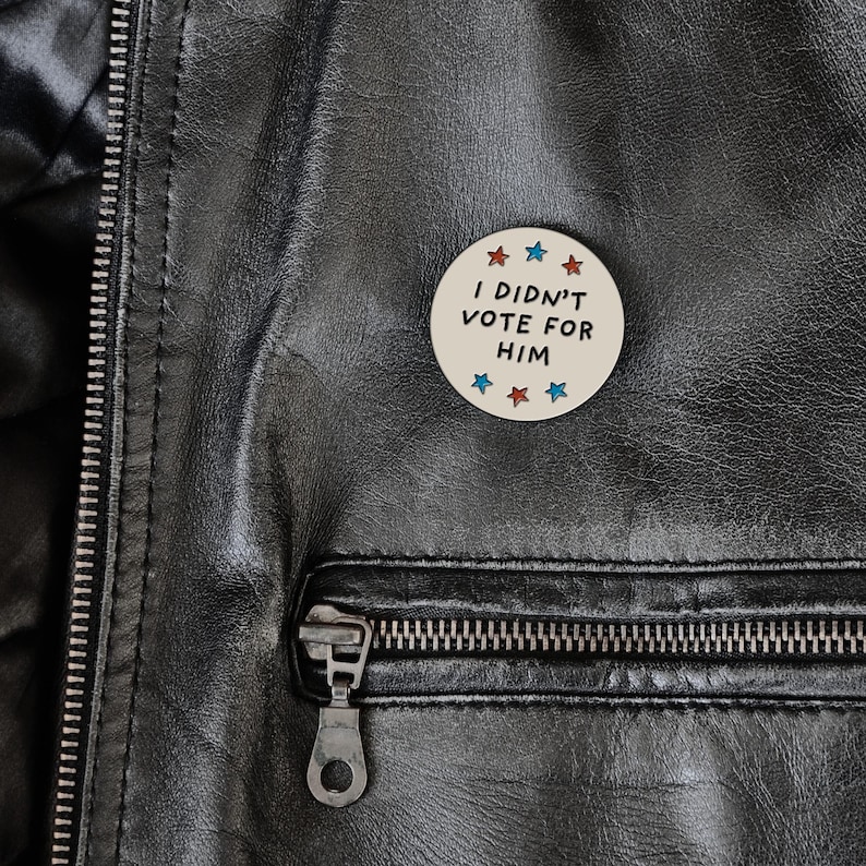May include: A round, off-white button with the text "I DIDN'T VOTE FOR HIM" in black. The button is decorated with red, white, and blue stars. The button is pinned to a black leather jacket.