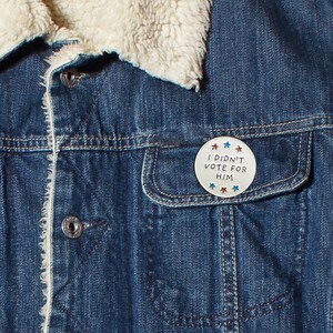May include: Close-up of a denim jacket with a white, faux-shearling collar. A round, white button is pinned to the pocket, featuring the text "I DIDN'T VOTE FOR HIM" with red and blue star accents. The jacket is a medium wash blue.