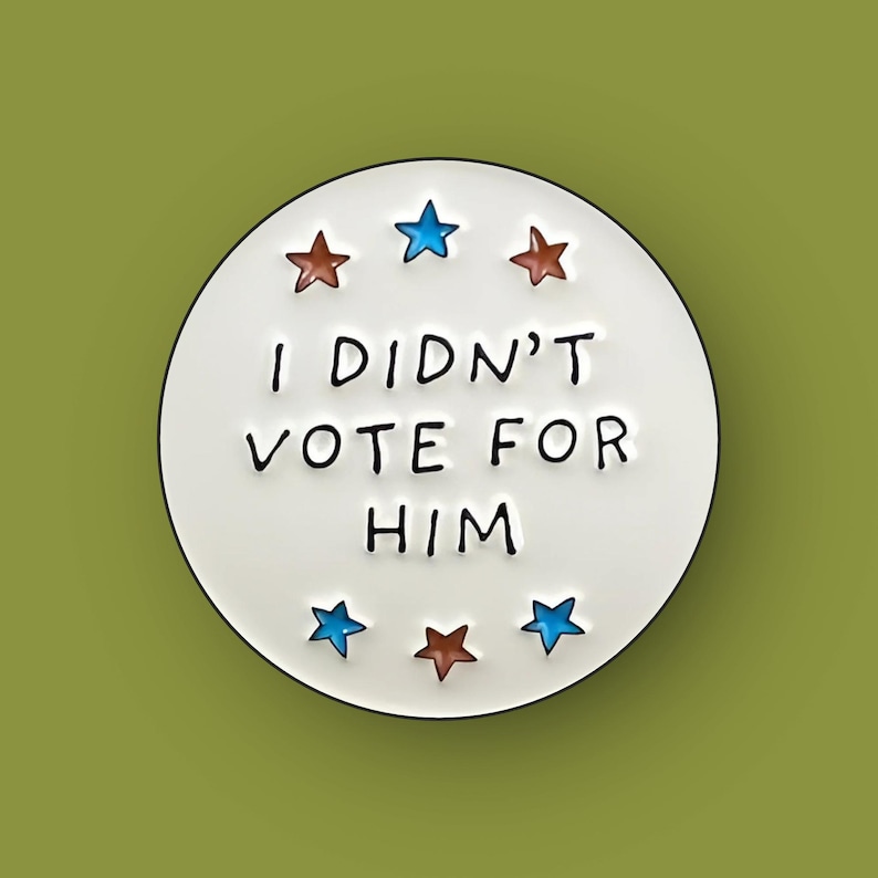 May include: A white circular pin with the text "I DIDN'T VOTE FOR HIM" in black. The pin is decorated with six stars, three brown and three blue, arranged around the text. The background is a solid olive green.