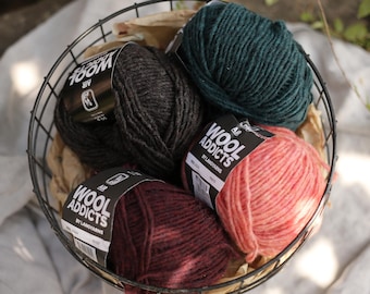 Op de afbeelding: Een metalen draadmand gevuld met bollen wol in verschillende kleuren, waaronder zwart, teal en koraal. Het garen is gemarkeerd met "Wool Addicts" en "100M Air". De mand staat op een lichtgekleurd oppervlak.
