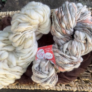 May include: Two large skeins of thick, textured yarn and one smaller skein rest in a woven basket. The left skein is cream-colored with a pink label reading Pixie Dust. The right skeins feature a blend of white, gray, and peach tones with a red label reading Oak.