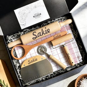 May include: A black gift box containing a wooden rolling pin, a wooden spoon, a dough docker, a metal dough scraper, and a red and white checkered kitchen towel. Each wooden item is engraved with the name Sakie. A card reads Fresh Bread Everyday.