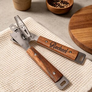 May include: A stainless steel can opener with wooden handles. The handles are engraved with the name "Rachael". The can opener is resting on a white waffle-weave cloth, with a small potted plant and wooden bowl in the background.