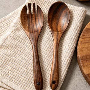 K&ouml;nnte beinhalten: Ein Salatbesteck aus Holz mit Gabel und L&ouml;ffel. Die dunkelbraunen Holzutensilien liegen auf einem cremefarbenen Waffelstoff. Die Gabel hat drei Zinken, und beide Teile haben ein Loch im Griff.