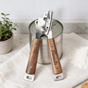 May include: A stainless steel can opener with wooden handles. The handles are engraved with the name "Fiametta". The can opener is positioned over a metal can, with a white cloth, flour, and cookies in the background.
